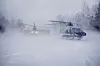 To politihelikoptre lander i kraftig snøfokk på en åpen, snødekt plass.