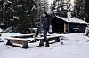 Mann står ved snødekt benk utenfor en liten hytte i vinterskog.