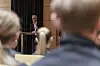 En mann står ved en talerstol og snakker til en gruppe mennesker i et møte- eller konferanserom.