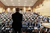 En person står foran en stor forsamling i et auditorie med mange tilhørere på rekke og rad.