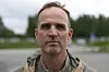 En mann står ute i dagslys foran en uskarp bakgrunn av trær og bygninger.