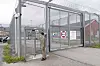 En mann står ved en åpen metallport inn til et inngjerdet område med piggtråd.