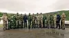 En gruppe personer i militæruniform står på rekke ute på en våt grusplass.