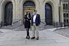 To personer står på en steinlagt plass foran en stor historisk bygning med buede dører og trapper.