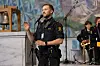 En mann i politiuniform taler ved mikrofoner foran en scene med musikere i bakgrunnen.