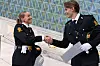 To personer i mørke uniformer håndhilser innendørs, den ene holder papirark.