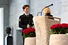 En uniformert person står ved en talerstol innendørs, med røde blomster og mikrofon i forgrunnen.