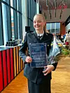 Smilende person i uniform holder diplom, blomster og trofé i en lys hall.