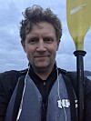 En mann med vindblåst hår holder en gul padleåre mot himmelen.