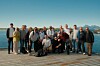 En gruppe mennesker står samlet utendørs på en brygge med sjø og fjell bak.