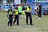 Tre politibetjenter i refleksvester går på en gressbane med fotballspillere i bakgrunnen.