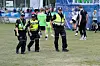 Tre politibetjenter i refleksvester går på en gressbane med fotballspillere i bakgrunnen.