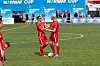 To fotballspillere i rødt står ved ballen på en gressbane med blå Norway Cup-bannere i bakgrunnen.