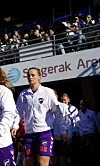 Fotballspillere i hvite jakker og lilla shorts går på banen foran en tribune med publikum.
