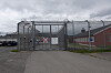 En metallport med piggtråd foran en grå bygning og et rødt murhus på en overskyet dag.