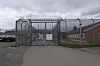 En metallport med piggtråd foran en grå bygning og et rødt murhus på en overskyet dag.
